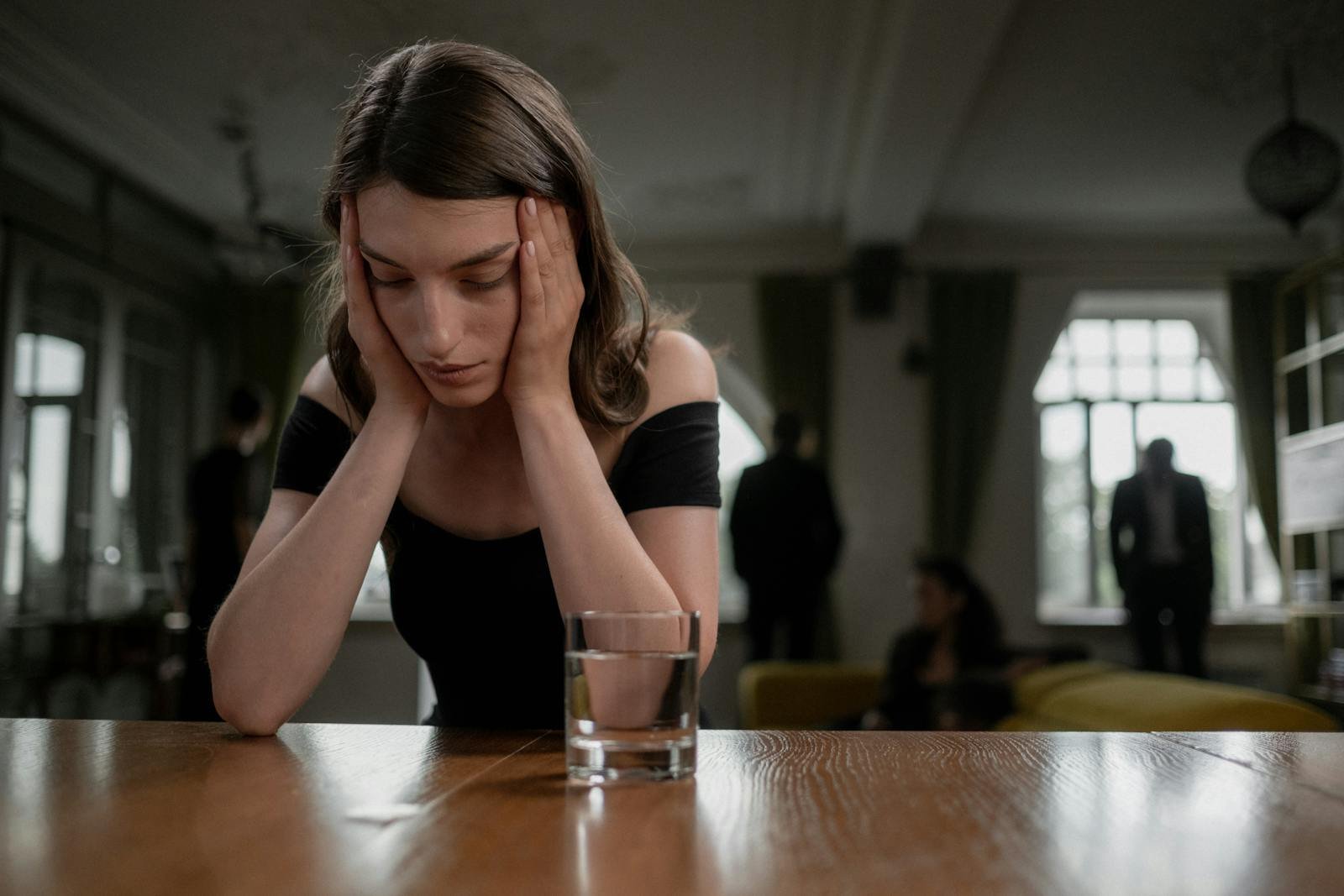 A sad woman in a black dress with a glass of water, indoors.